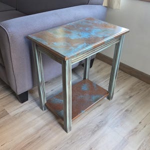 Pode incluir: Uma pequena mesa lateral com um tampo retangular e uma prateleira inferior. O tampo tem um acabamento envelhecido com tons de azul, castanho e cobre. A estrutura e as pernas são pintadas em um tom verde-azulado suave. A mesa está ao lado de um sofá cinza.