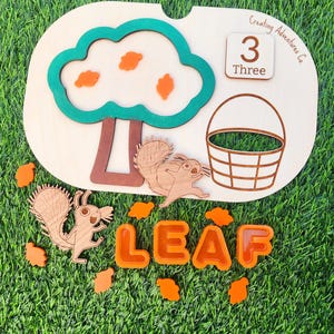 May include: Wooden educational toy featuring a tree with orange leaves, two squirrels, a basket, and the word "LEAF" spelled out in orange letters. The number "3" and the word "Three" are also included on the toy.