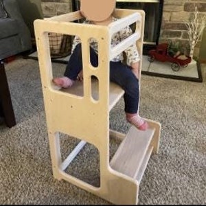 May include: A wooden learning tower with two steps and a safety rail. The tower is made of natural wood and has a light brown finish. A child is sitting on the top step of the tower.
