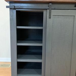 May include: A gray cabinet with a sliding door and shelves. The cabinet has a dark wood top and black hardware.