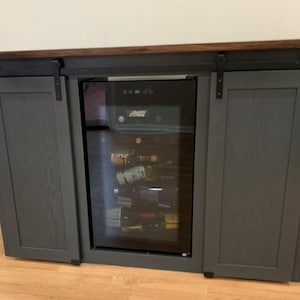 May include: A gray wooden cabinet with sliding barn doors and a built-in wine cooler. The cabinet has a dark wood countertop and black metal hardware.