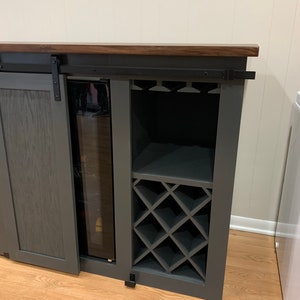 May include: A dark gray wooden bar cabinet with a sliding door, a built-in wine rack, and a small refrigerator. The cabinet has a dark wood top and black metal hardware.