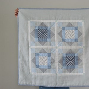Op de afbeelding: Een lichtblauwe en grijze quilt met een geometrisch patroon. De quilt heeft vier grote vierkanten, elk met een centraal ontwerp in tinten blauw en wit. De rand is lichtblauw. De quilt wordt door onzichtbare handen omhooggehouden.