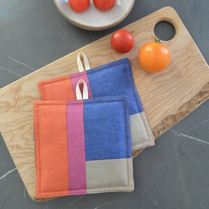 Puede incluir: Dos salvamanteles cuadrados y coloridos con un lazo para colgar. Cada salvamanteles tiene tres secciones verticales en naranja, rosa y azul. Una tabla de cortar de madera y tomates en el fondo.