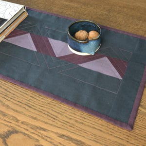 May include: A rectangular, dark teal table runner with geometric patterns in shades of purple and burgundy. A small, blue ceramic bowl with two walnuts sits on the runner. A book with a gold-colored metal disc is on the left.