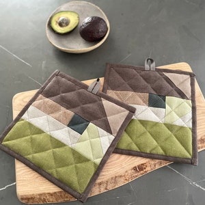 Op de afbeelding: Twee vierkante, gewatteerde pannenlappen met geometrische patronen in bruin-, groen- en beigetinten. Elke pannenlap heeft een lus om op te hangen. De pannenlappen liggen op een houten snijplank, met een kom met avocado's op de achtergrond.