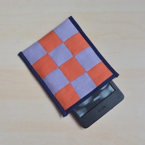 Peut inclure: Une housse faite à la main pour liseuse Kindle avec un motif en damier orange et lavande. L'étui a une bordure bleu marine et est partiellement ouvert, révélant l'appareil Kindle. L'étui est en tissu.
