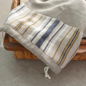 Può includere: Una borsa di tessuto a righe con chiusura a coulisse, realizzata in lino naturale con strisce marroni e blu. La borsa è in un cesto di vimini.