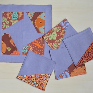 Może przedstawiać: Zestaw ręcznie robionych podkładek i kwadratowej podkładki. Podkładki i podkładka są lawendowe z trójkątami z wzorzystej tkaniny w odcieniach brązu, pomarańczy i turkusu. Podkładka ma wymiary około 20 cm kwadratowych.