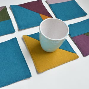 Może przedstawiać: Zestaw kolorowych podkładek z tkaniny o geometrycznych wzorach. Każda podkładka ma kombinację turkusu, fioletu, zieleni i żółci. Jasnoniebieski ceramiczny kubek stoi na jednej z podkładek. Podkładki są kwadratowe.