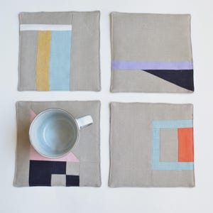 Może przedstawiać: Cztery kwadratowe szare lniane podstawki z geometrycznymi wzorami bloków kolorów. Jedna podstawka ma jasno-niebieski i żółty pasek, druga fioletowy pasek i czarny trójkąt, trzecia różowy trójkąt i granatowy kwadrat, a ostatnia niebieski i pomarańczowy kwadrat.
