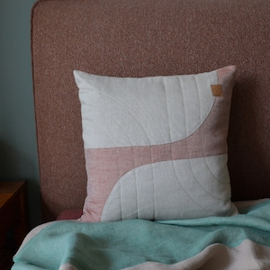 Può includere: Cuscino quadrato decorativo con un design grigio chiaro e rosa antico. Il cuscino presenta un motivo trapuntato con una forma curva e astratta. Una piccola etichetta quadrata in pelle è attaccata all'angolo in alto a destra. Il cuscino è appoggiato su un letto con una coperta verde acqua.