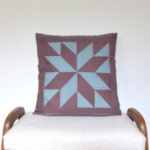 May include: A square decorative pillow with a starburst pattern. The pillow features a central star design in light blue and a dark mauve background. The pillow is placed on a white fluffy surface.