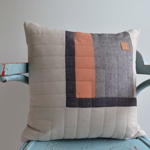 Può includere: Cuscino quadrato decorativo con un design patchwork. Il cuscino presenta uno sfondo trapuntato grigio chiaro con un design quadrato grigio scuro, arancione e grigio. È attaccata una piccola etichetta in pelle.