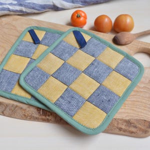 Può includere: Due presine quadrate con un motivo a scacchiera di quadrati gialli e blu-grigi. Ogni presina ha un bordo verde e un anello blu per appenderla. Le presine sono su un tagliere di legno con pomodori e un cucchiaio di legno.