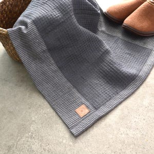 Puede incluir: Una manta gris con tejido de gofre, borde a rayas y una pequeña etiqueta de cuero. La manta está doblada cerca de una cesta de mimbre y un par de zapatillas de ante marrón. La textura y el color sugieren comodidad y relajación.