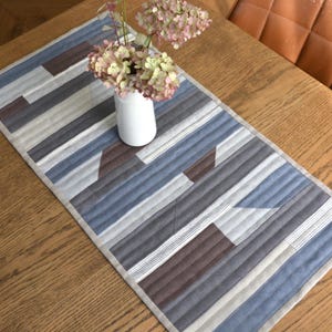 Può includere: Runner da tavolo rettangolare trapuntato con un motivo geometrico nei toni del blu, grigio e marrone. Il runner ha un bordo beige chiaro ed è posto su un tavolo di legno. Un vaso bianco con fiori rosa è sopra.