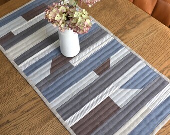 May include: A rectangular quilted table runner with a geometric pattern in shades of blue, gray, and brown. The runner has a light beige border and is placed on a wooden table. A white vase with pink flowers sits on top.
