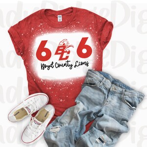 May include: Red t-shirt with a white bleach design featuring the numbers 666, the letters "BC" and the text "Boyd County Lions". The shirt is on a white background with a pair of white sneakers and blue jeans.