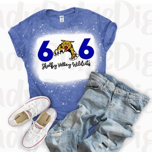 May include: A blue t-shirt with the number "66" in navy blue and a wildcat graphic. The text "Shelby Valley Wildcats" is below the graphic. The shirt is paired with light-wash jeans and white sneakers.