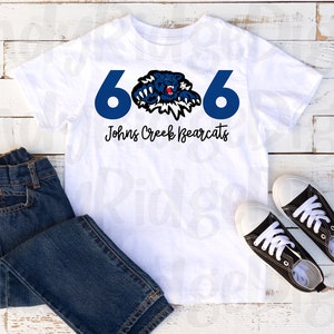 May include: White t-shirt with a blue number 6 on each side and a black bear mascot in the center. The text "Johns Creek Bearcats" is printed below the mascot.