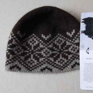 May include: A brown and gray knitted hat with a geometric pattern. The hat is made of yak down and has a tag that reads "Yak Down" with information about the material and its origin. Made in Mongolia.