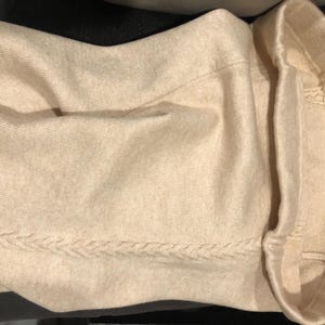 May include: A light beige sweater with a braided design along the bottom hem. The sweater has a ribbed collar and a small tag on the cuff. The fabric appears soft and comfortable.