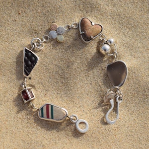 Op de afbeelding: Een zilveren armband met verschillende bedels, waaronder een hartvormige steen, een kleine rood-wit gestreepte steen en een kleine zwarte steen. De armband ligt op een zanderig oppervlak.