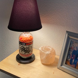 May include: A table lamp made from a repurposed tequila bottle filled with orange lentils. The bottle has a black and white label with the text "Puro Agave Espolon". The lamp has a dark purple lampshade.