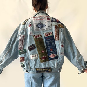 May include: A light blue denim jacket featuring numerous rectangular patches with various text and graphic designs across the back. The jacket has a classic collar, long sleeves, and a button-up front, paired with matching light blue denim pants.
