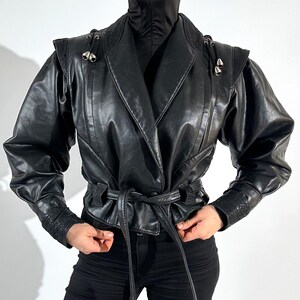 May include: Black leather jacket with a wrap design, featuring a notched collar, shoulder detailing, and a tie belt. The jacket has long sleeves with detailed cuffs and is paired with black trousers. Silver-toned hardware accents the shoulders.