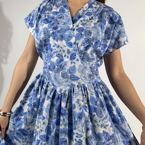 May include: A vintage blue floral dress with a V-neckline and short sleeves. The dress features a full skirt and a fitted waist. The floral pattern includes shades of blue and white. The dress is made of a lightweight fabric.