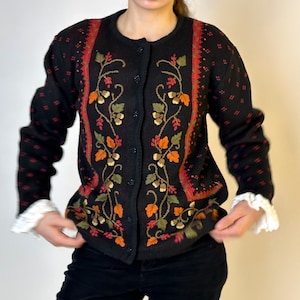 May include: Black button-up cardigan sweater with an embroidered floral design in orange, green, and gold. The sweater has red accents and white lace cuffs. The overall theme is vintage apparel.
