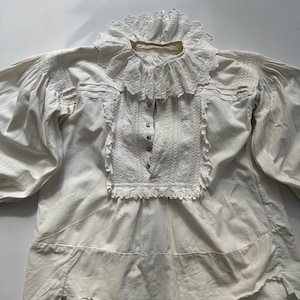 May include: A white cotton blouse with a lace collar and trim. The blouse has a gathered neckline and long sleeves. The lace is delicate and intricate.