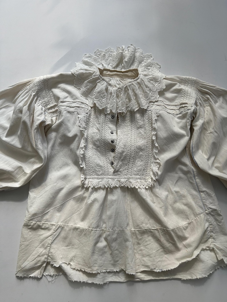 May include: A white cotton blouse with a lace collar and trim. The blouse has a gathered neckline and long sleeves. The lace is delicate and intricate.