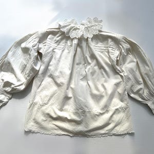 May include: A white cotton blouse with a lace collar and long sleeves. The blouse has a gathered neckline and a slightly ruffled hem.