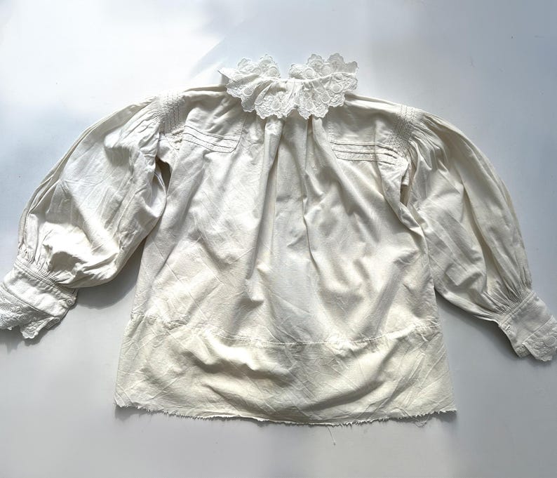 May include: A white cotton blouse with a lace collar and long sleeves. The blouse has a gathered neckline and a slightly ruffled hem.