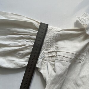 May include: A white cotton blouse with a gathered detail at the shoulder and a lace trim at the neckline. The blouse is measured with a ruler in inches.