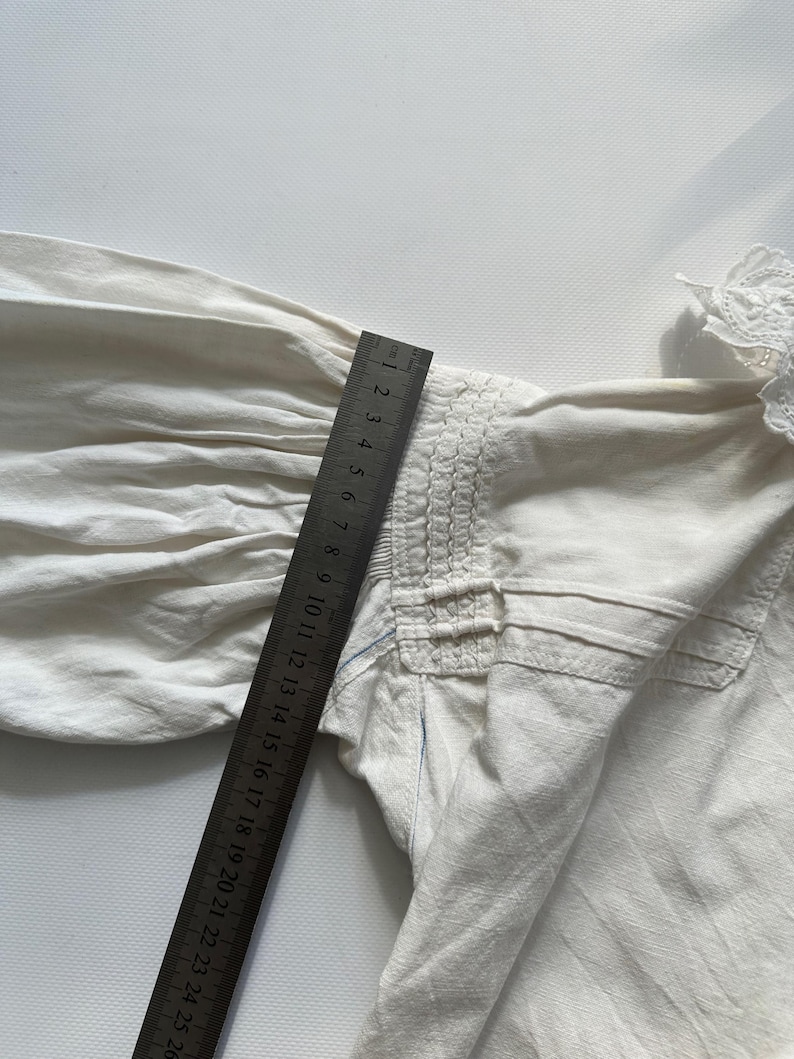May include: A white cotton blouse with a gathered detail at the shoulder and a lace trim at the neckline. The blouse is measured with a ruler in inches.
