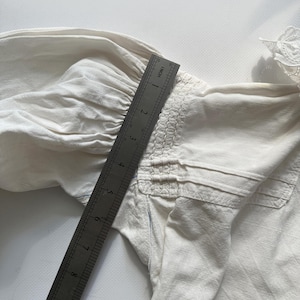 May include: A white cotton blouse with a gathered detail at the shoulder and a lace trim at the neckline. A ruler is placed on the blouse to show the size.