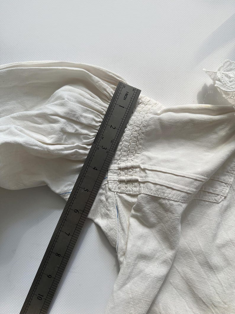 May include: A white cotton blouse with a gathered detail at the shoulder and a lace trim at the neckline. A ruler is placed on the blouse to show the size.