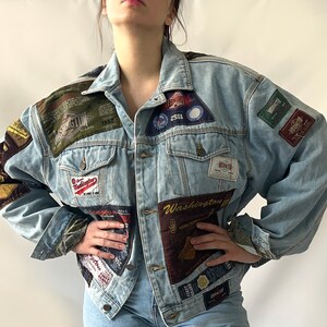 以下が含まれることがあります： 様々なパッチが縫い付けられた薄いブルーのデニムジャケット。パッチは異なる生地で作られており、テキスト、ロゴ、画像など、様々なデザインが施されています。ジャケットはボタン留めのフロントと2つのフラップポケットを持つ、クラシックなデニムスタイルです。