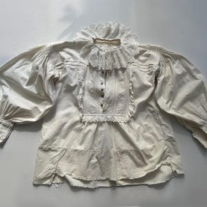 May include: A white cotton blouse with a lace collar and long sleeves. The blouse has a gathered neckline and a ruffled hem.