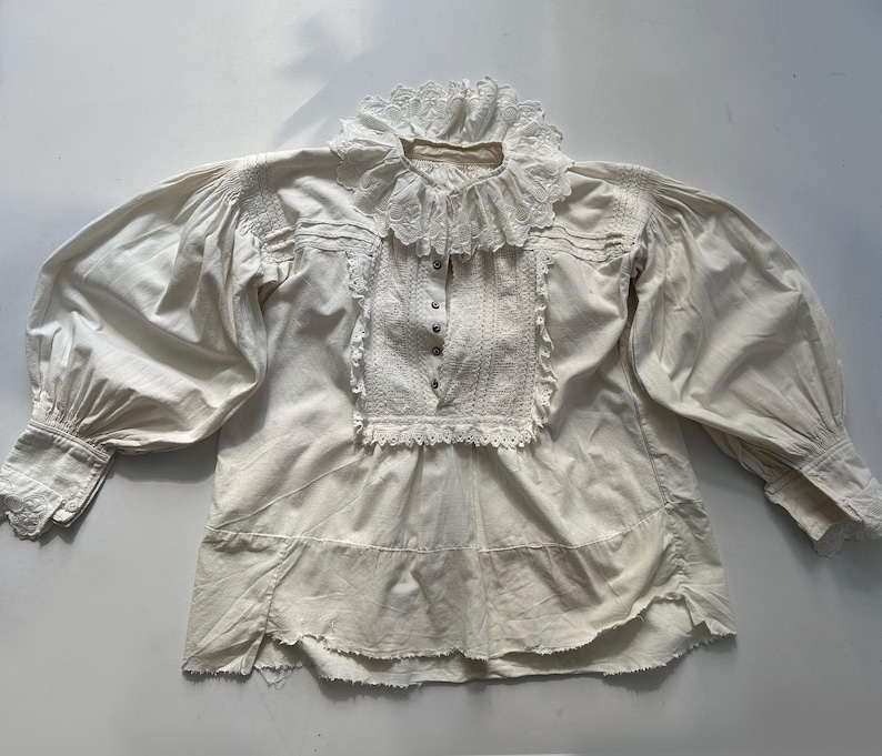 May include: A white cotton blouse with a lace collar and long sleeves. The blouse has a gathered neckline and a ruffled hem.