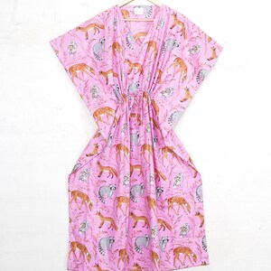 May include: A pink, V-neck caftan dress with a drawstring waist. The dress features a repeating pattern of foxes, deer, raccoons, and floral elements. The caftan is hanging on a wooden hanger against a white background.