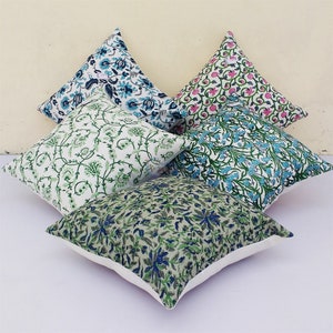 Puede incluir: Cuatro almohadas cuadradas con patrones florales en tonos verdes, azules y rosas sobre un fondo blanco. Las almohadas están apiladas.