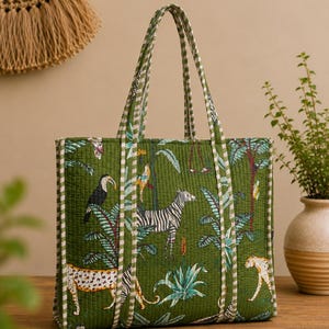 May include: A quilted tote bag in a deep olive green fabric featuring a vibrant jungle print with illustrations of a zebra, a leopard, and a toucan among tropical plants. The bag includes two long shoulder straps with a white and green striped pattern.