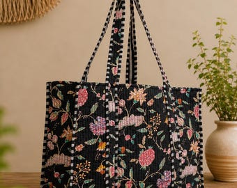 Op de afbeelding: Een gewatteerde tote bag met een zwarte basis en een veelkleurig bloemenpatroon in roze, oranje en paarse tinten. De tas heeft twee lange hengsels met een zwart-wit gestreepte bies die past bij de afwerking langs de randen van de tas.