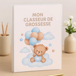 Może przedstawiać: Przezroczysty segregator z jasnoróżową okładką z ilustracją misia trzymającego balony. U góry wydrukowano tekst "MON CLASSEUR DE GROSSESSE". Obraz zawiera niebieskie i brązowe balony, chmury i gwiazdy. Złoty długopis leży na stole.