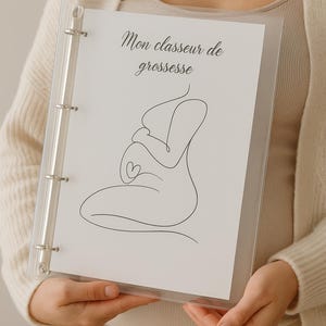 Peut inclure: Un classeur transparent avec une page blanche ornée d'un dessin au trait d'une femme enceinte et du texte "Mon classeur de grossesse". Le classeur a des anneaux métalliques argentés.
